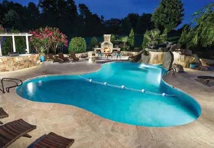 An illuminated, freeform swimming pool with a stone waterfall feature, surrounded by a paved patio and outdoor furniture.