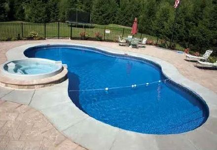 A kidney-shaped swimming pool with an attached circular spa, surrounded by stone decking, trees, and patio furniture.