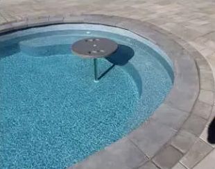 A round, gray table stands in a blue swimming pool, featuring a central support post resting on an underwater step.