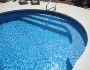 A blue circular swimming pool with built-in entry steps and a handrail, set against a light-colored concrete deck.