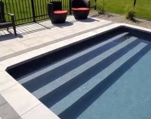 A backyard swimming pool with dark blue water and light stone stairs, next to a paved patio with two outdoor chairs.