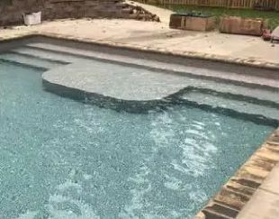 A light blue swimming pool with tiled steps and a built-in sun shelf, surrounded by a light-colored concrete deck.