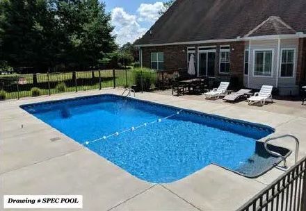 A rectangular blue swimming pool with a step entry area on a concrete patio beside a brick house on a sunny day.