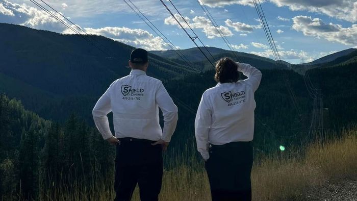 Two people in white sweatshirts stand on a mountain overlook facing a sunlit forested valley.