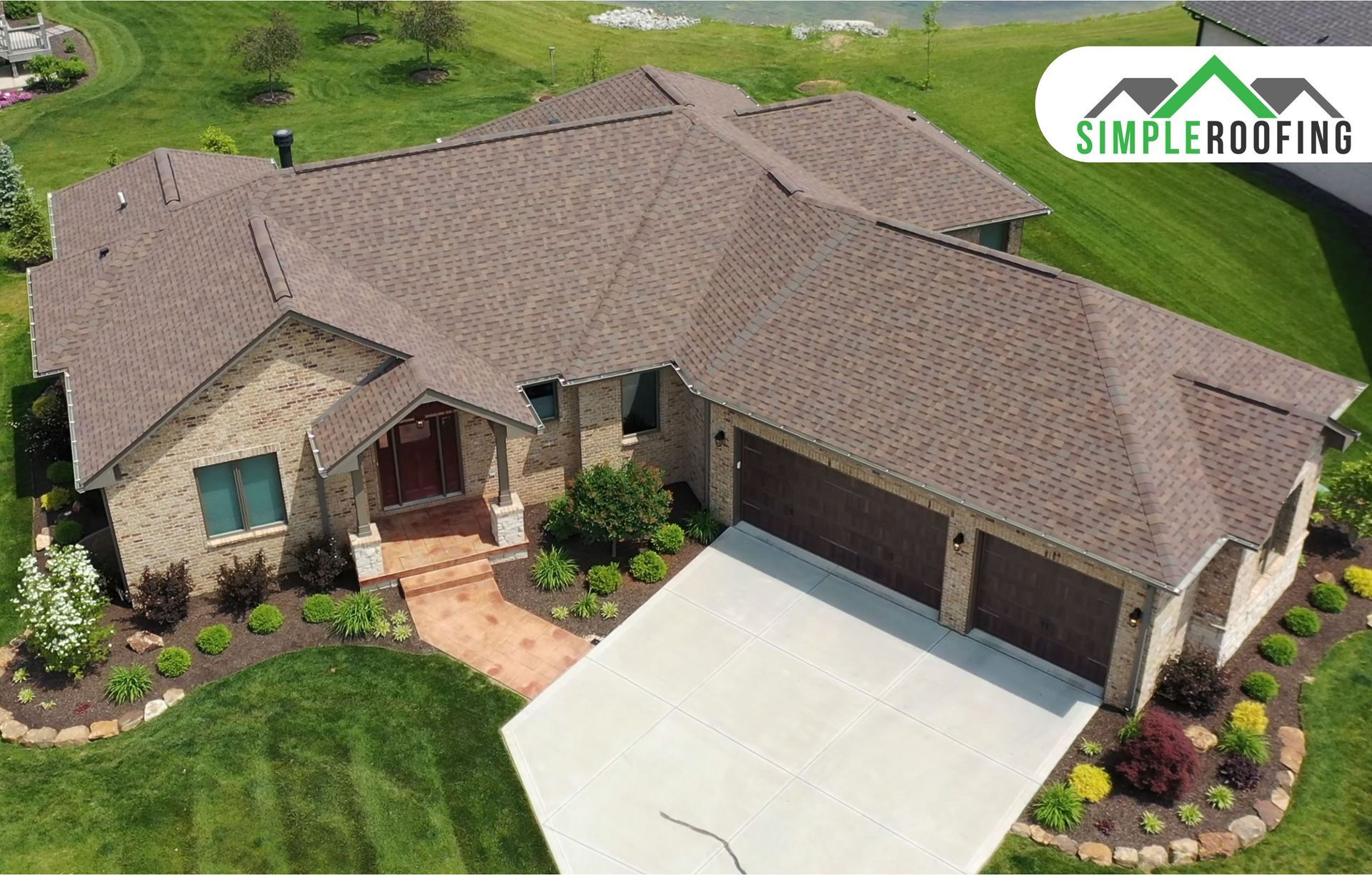 Aerial view of a suburban brick house with a brown shingled roof, a two-car garage, and a landscaped front yard.