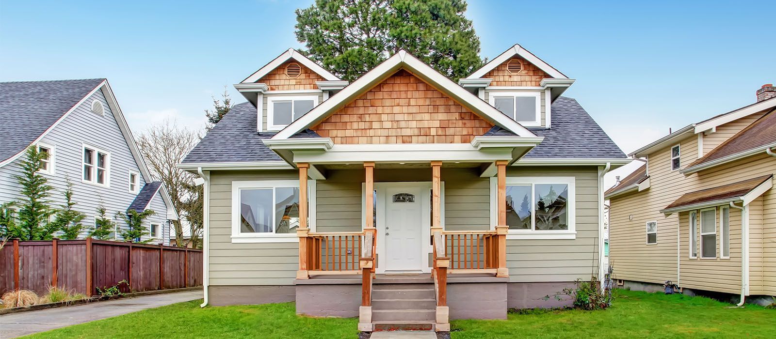 A beige house with a front porch, cedar shake gables, and two dormer windows, set between two other houses on a lawn.