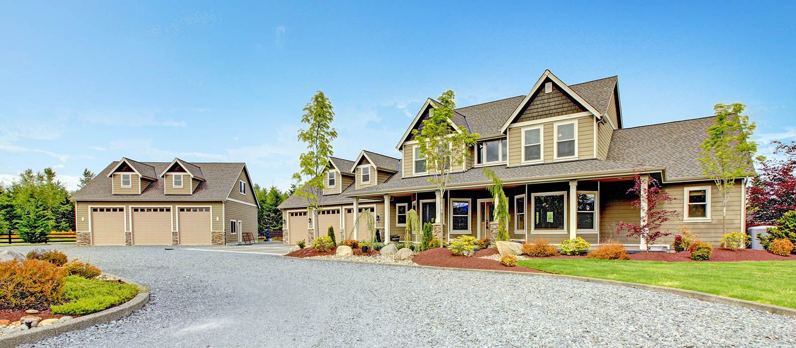A large tan-colored suburban home with a three-car garage and a gravel driveway under a bright blue sky.