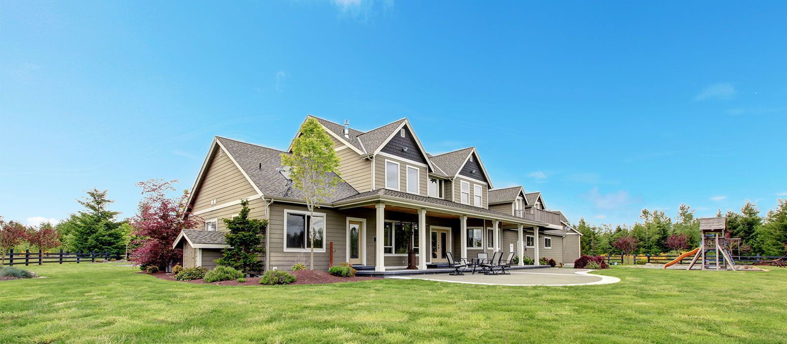 A multi-story house with tan siding and a stone patio set, surrounded by a green lawn under a clear blue sky.