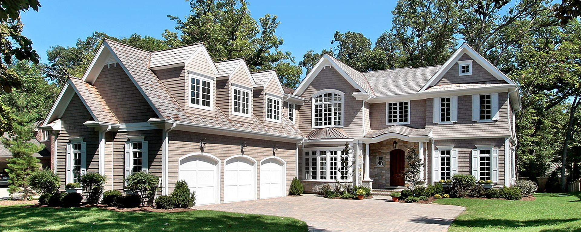 A large, two-story, light-brown house with a three-car garage, white trim, and dormer windows, surrounded by trees.