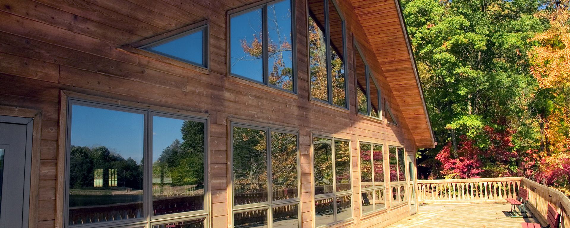 A wooden cabin with large, reflective windows, a deck, and surrounding autumn trees under a clear blue sky.