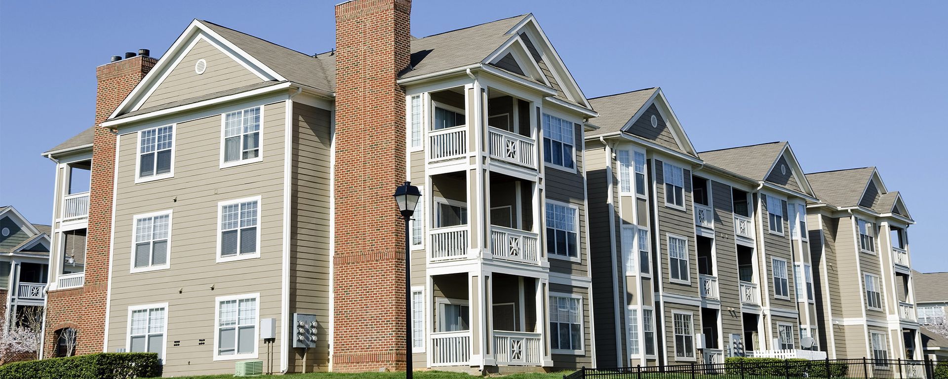 A multi-story apartment building with beige siding, brick chimneys, and white-trimmed balconies under a blue sky.