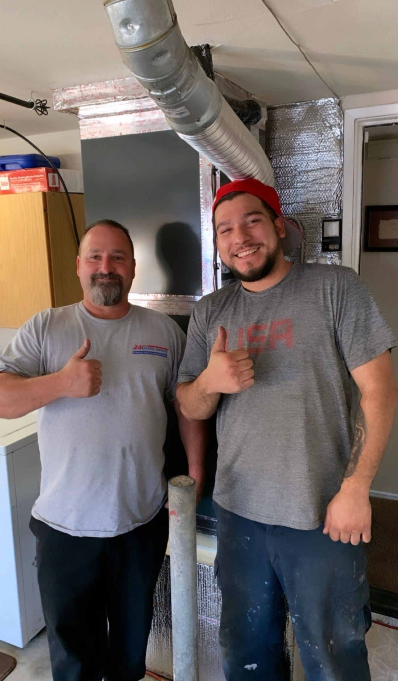 Two men giving thumbs-up next to installed HVAC system, indoors. One man wears a red hat.