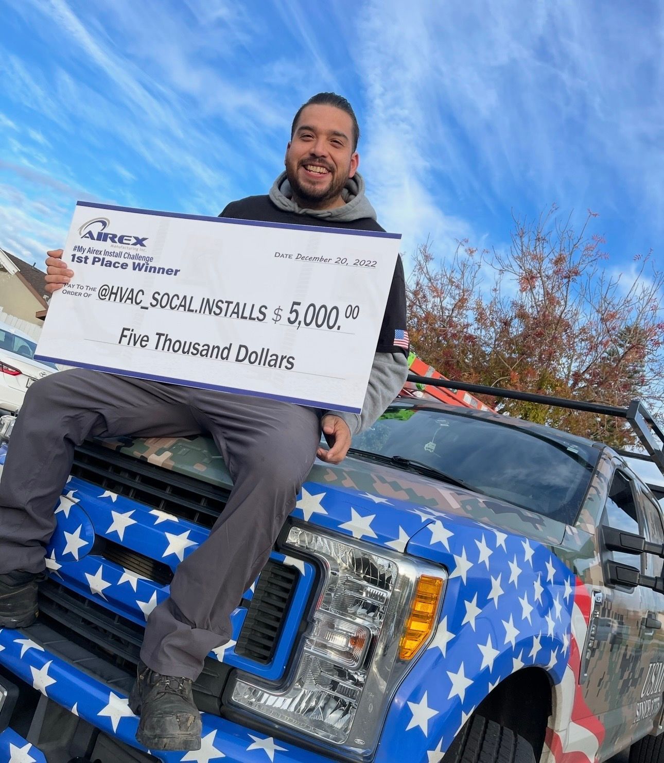 Man holding a check for $5,000, sitting on an American flag-themed truck, smiling against a cloudy sky.