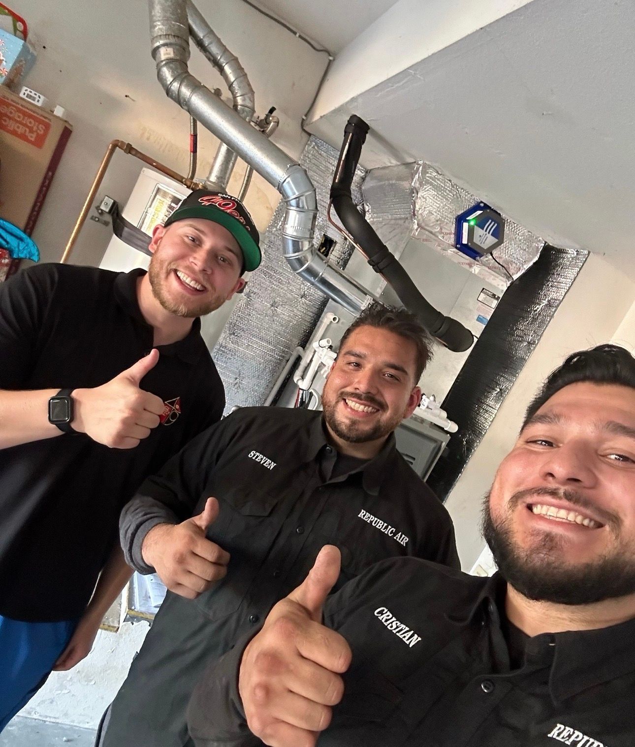 Three men in work uniforms give thumbs up in a garage near ductwork.