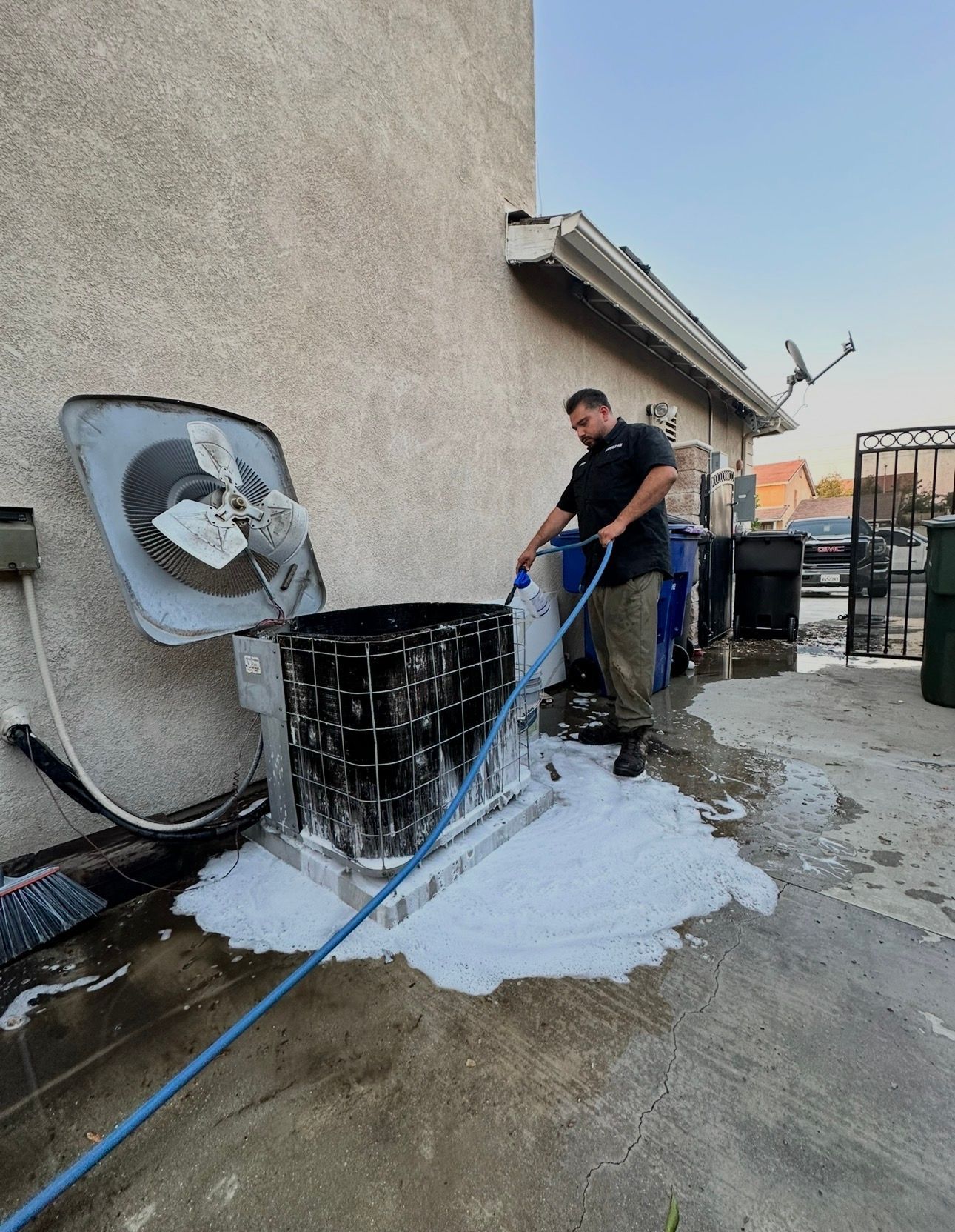 A man washes an air conditioner unit with soap and a hose outdoors, near a building.