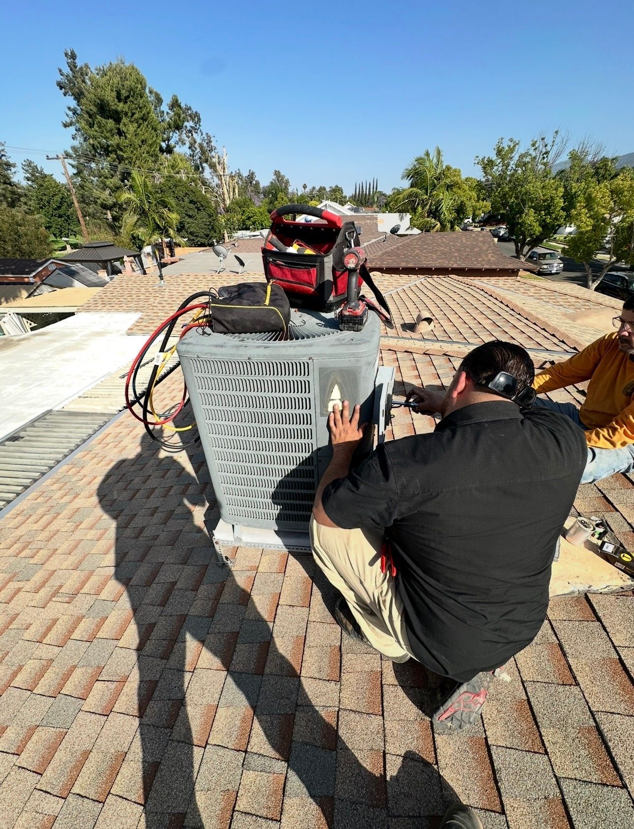 Two people working on an AC unit on a rooftop. One kneels, inspecting the unit; the other stands nearby.