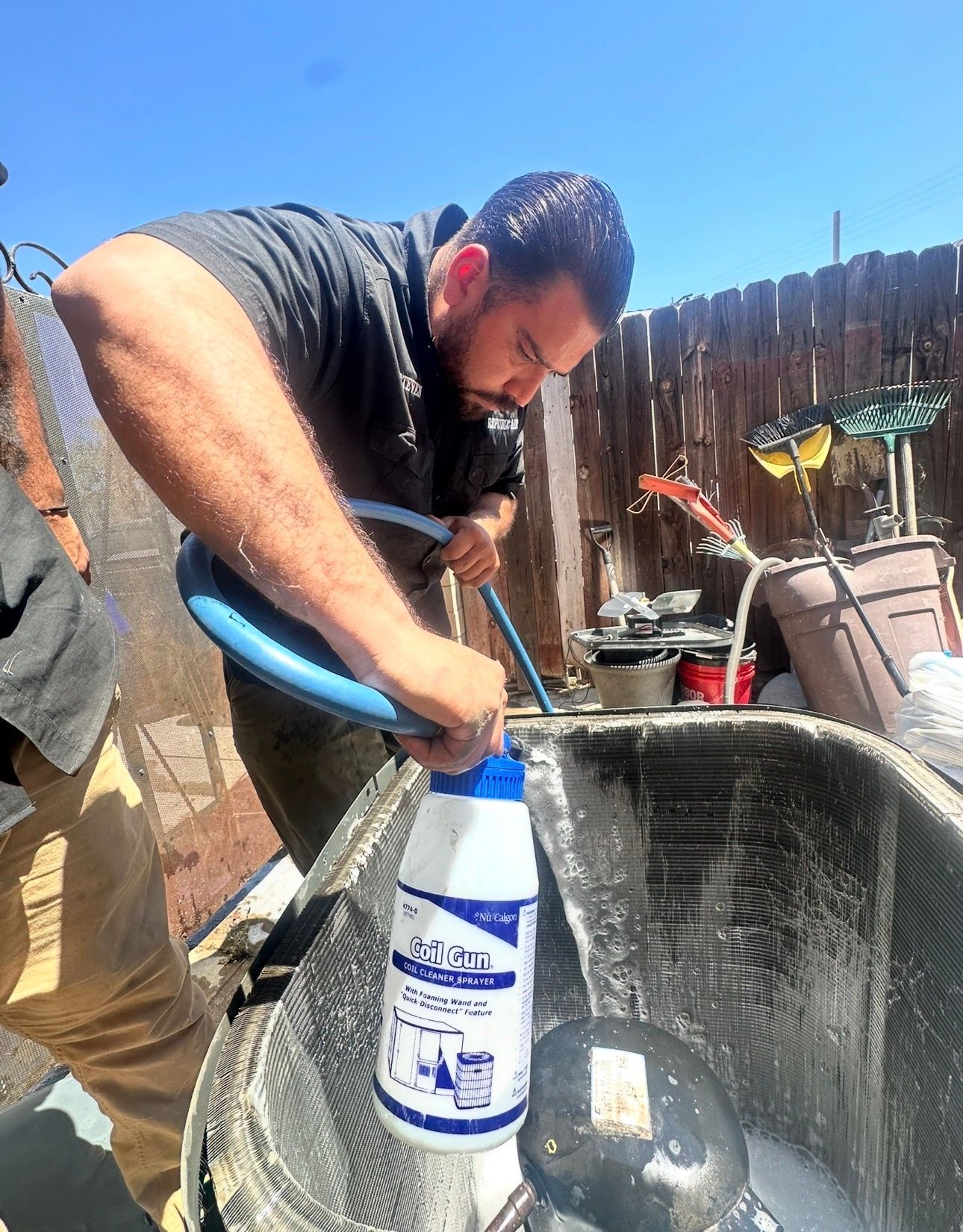 Man pouring liquid from a bottle into an air conditioning unit. Outdoors, sunny.
