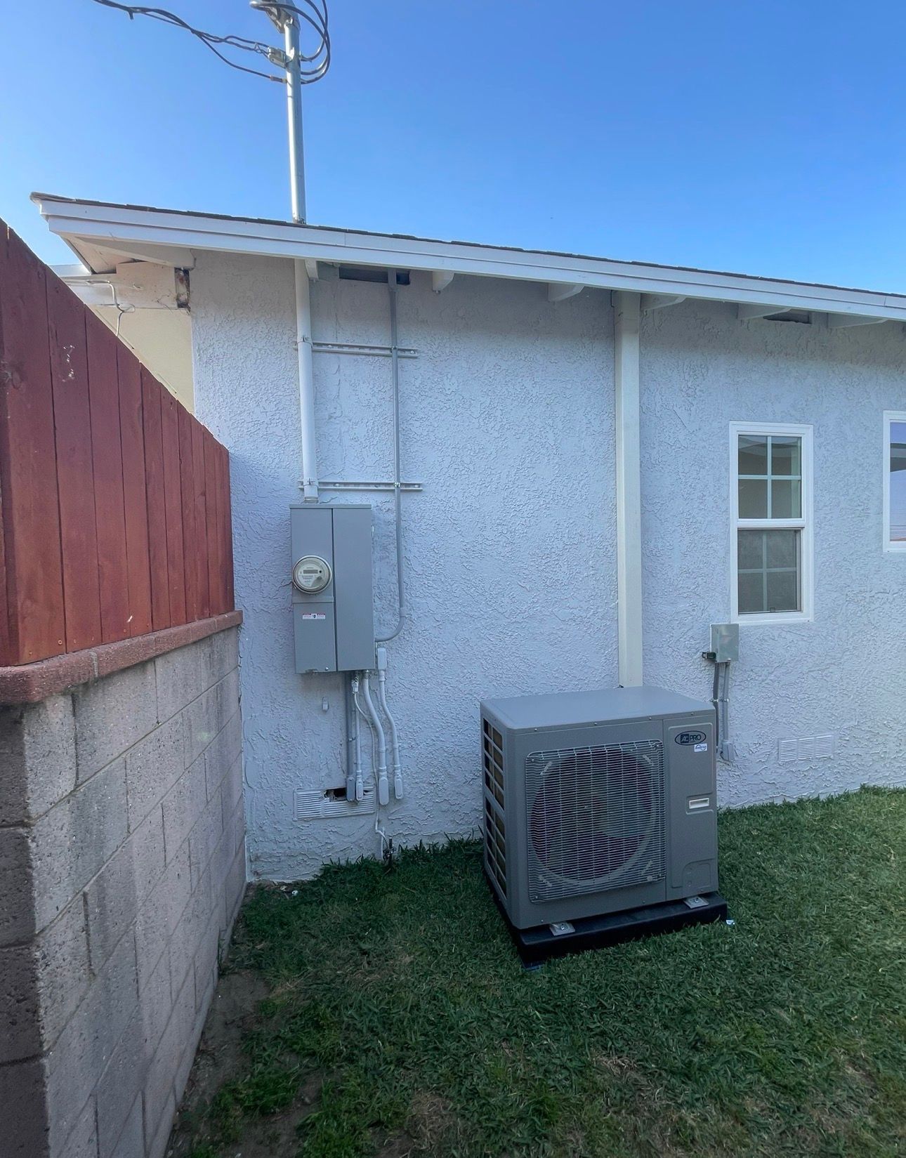 Outdoor electrical panel, AC unit, and building exterior. Gray panel and AC unit, white textured wall, green grass.
