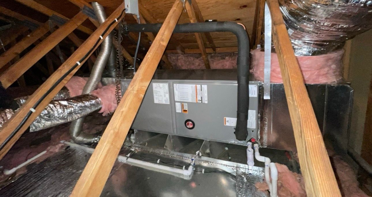 HVAC system in an attic. Gray metal unit with black pipes, surrounded by insulation and wooden beams.