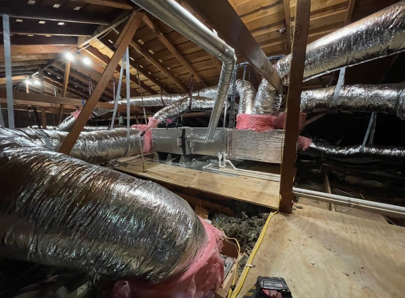 Attic with HVAC ductwork; insulation, wooden rafters, and plywood flooring visible.
