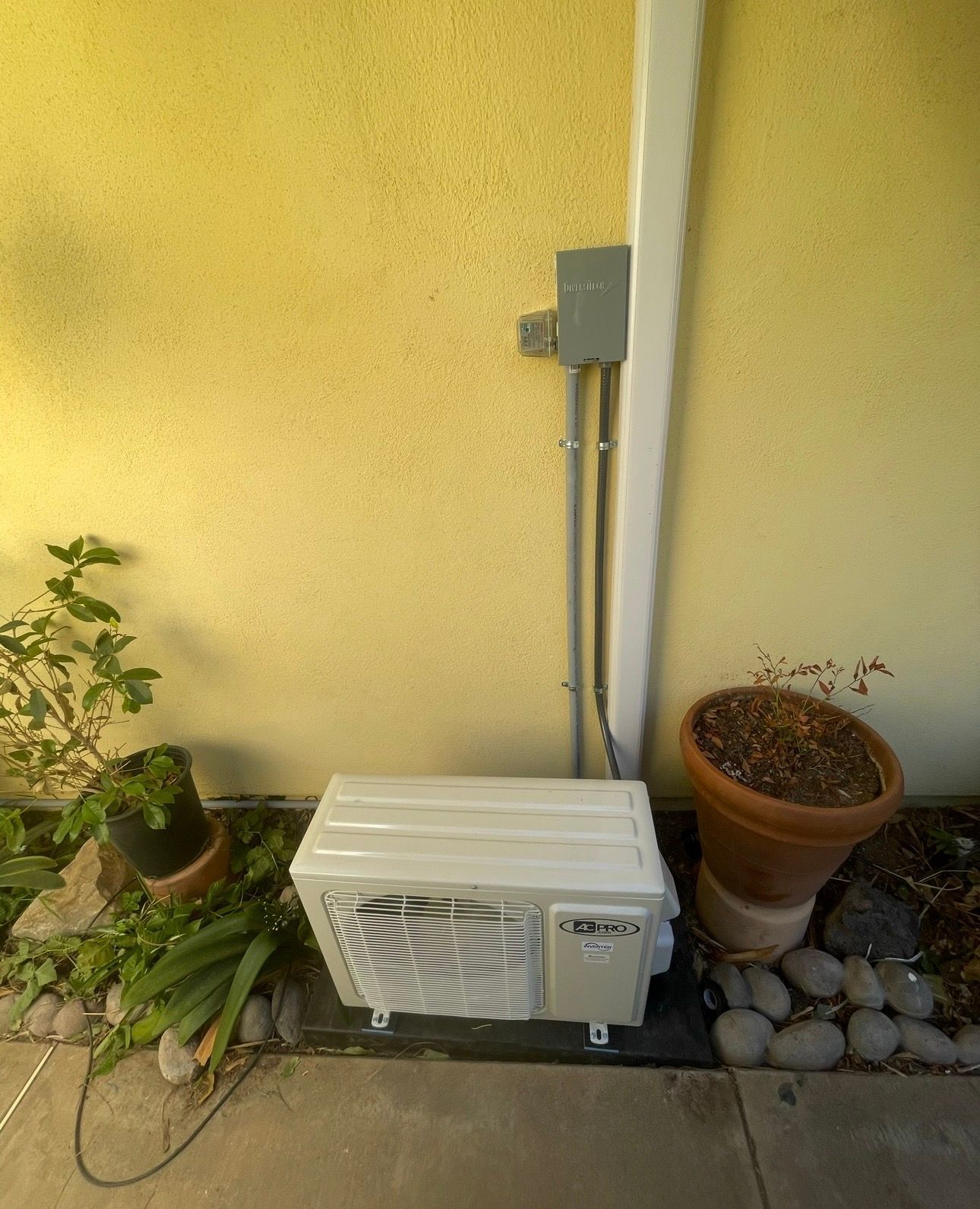 An air conditioning unit on a patio, next to a potted plant, rocks, and a yellow wall with electrical box.