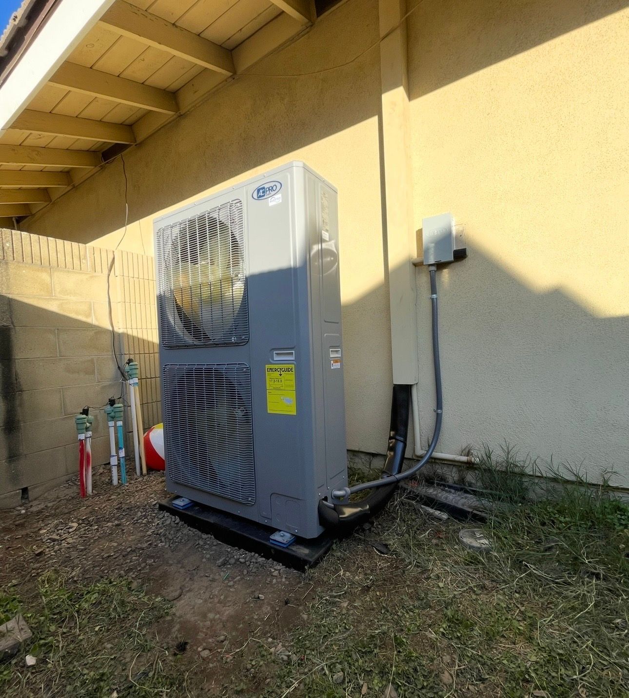 Gray heat pump unit next to a light-colored building with visible conduit and electrical box.