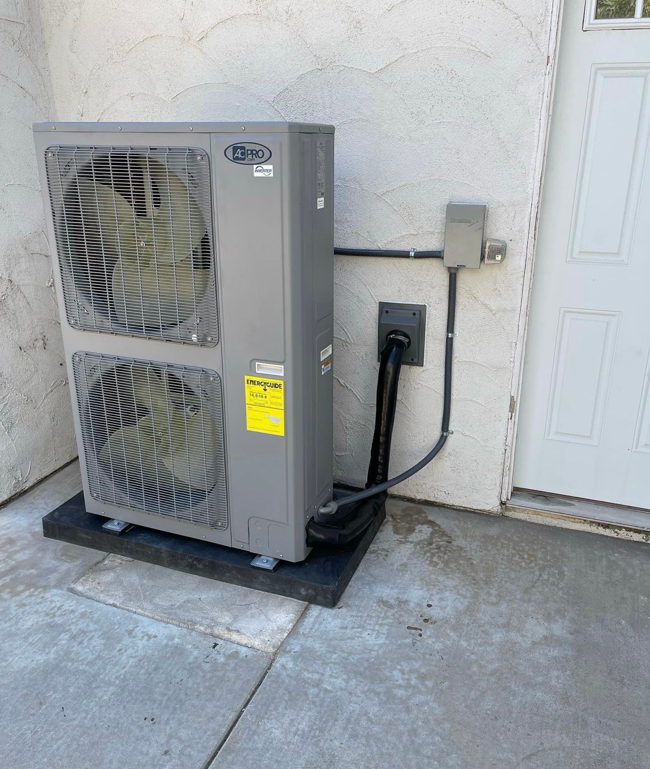 Gray heat pump unit next to a white wall and a door. Black electrical conduit and pad.