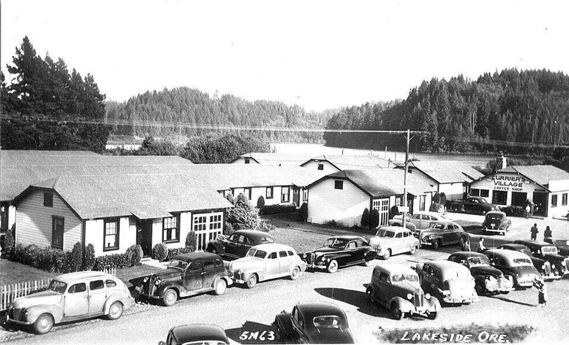 Vintage photo of Lakeside, Oregon with cars parked in front of several buildings, trees, and a lake in the background.