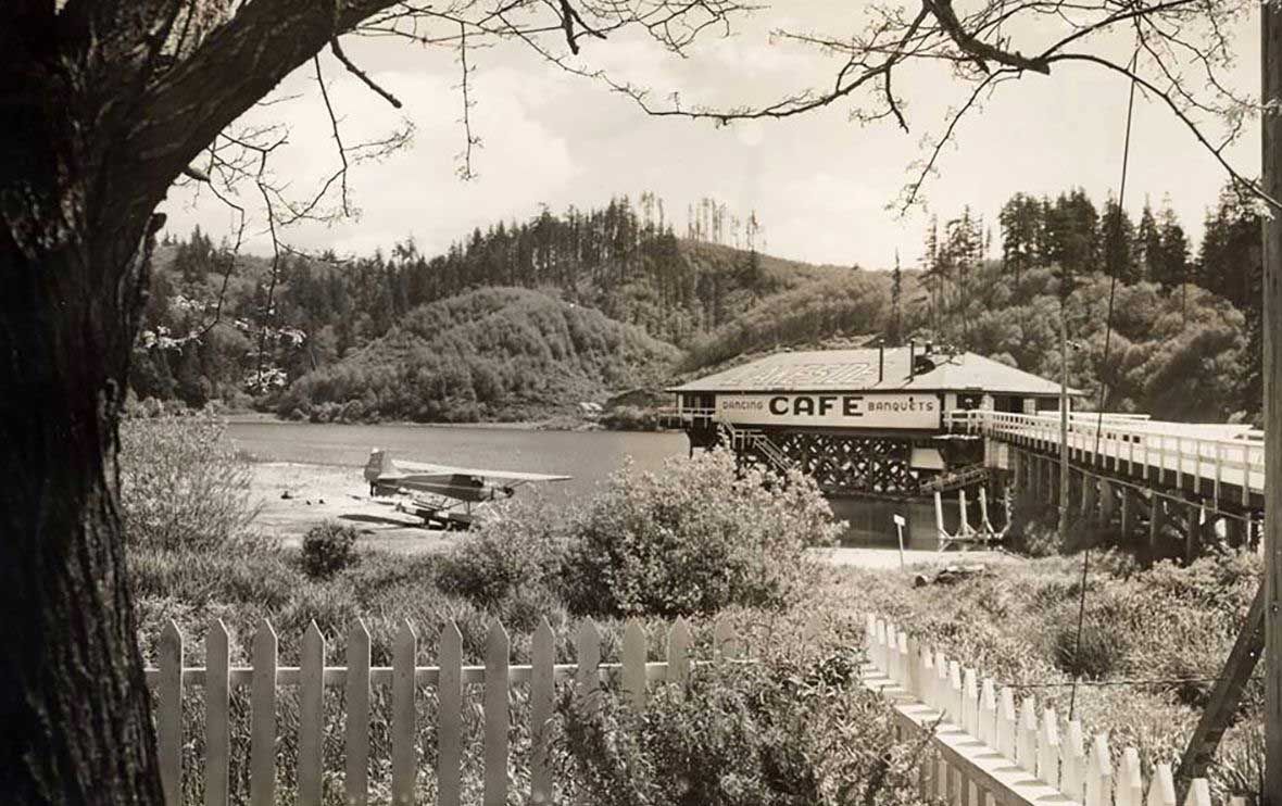 black and white photo of a cafe on a pier, a lake, and wooded background
