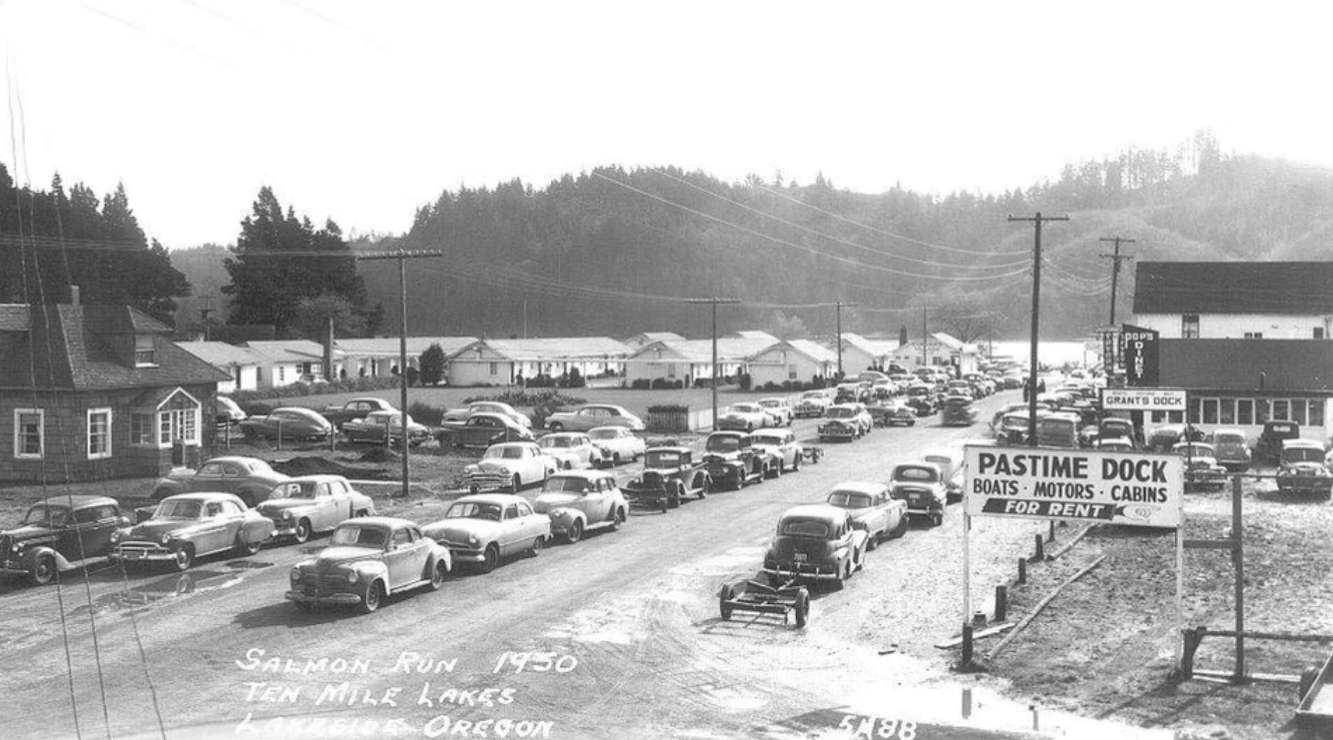 cars line a road near buildings and a dock in the 1950s