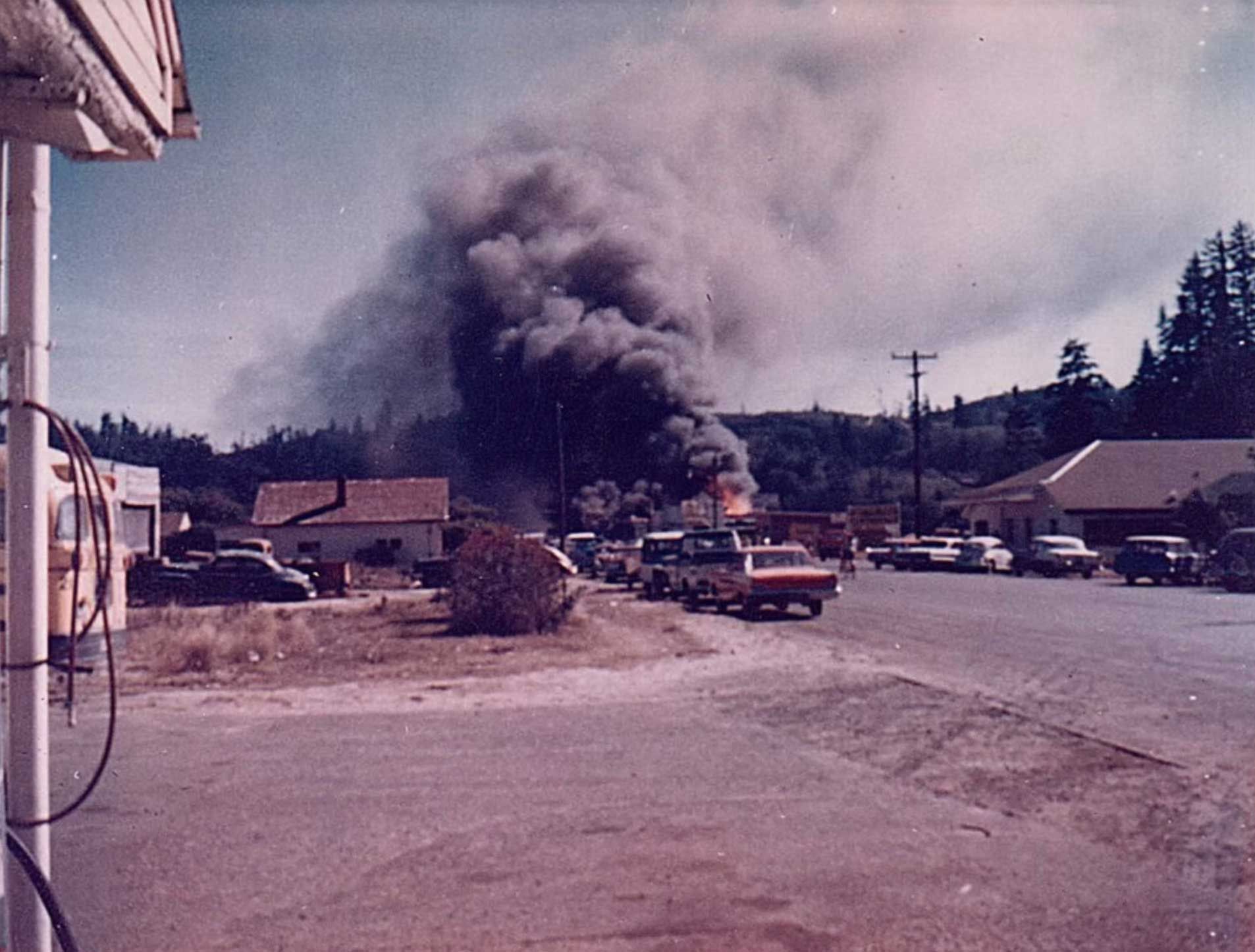 smoke billows over a town; a fire rages in the background, with cars and buildings in front