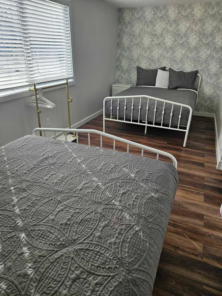 Two beds in a bedroom with patterned wallpaper. The beds have gray quilts, and one is closest to the viewer.