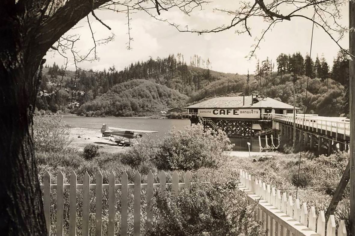 Black and white photo of seaplane at pier with cafe, trees in the background, behind a white picket fence.