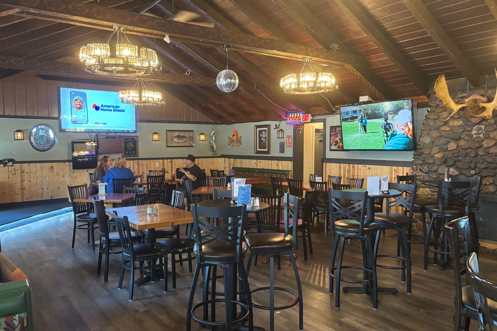 Interior view of a bar/restaurant with tables, chairs, TVs, and decorative lights. People are seated and dining.