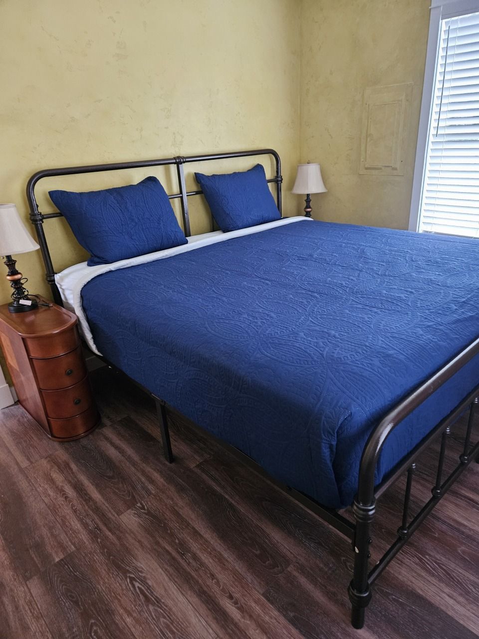 Bedroom with a dark metal bed frame, blue bedding, nightstands, and a window with blinds.