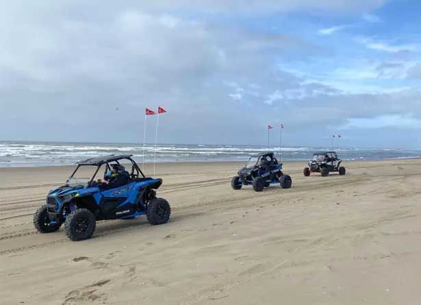 three off-road vehicles driving on a beach near the ocean, under a cloudy sky
