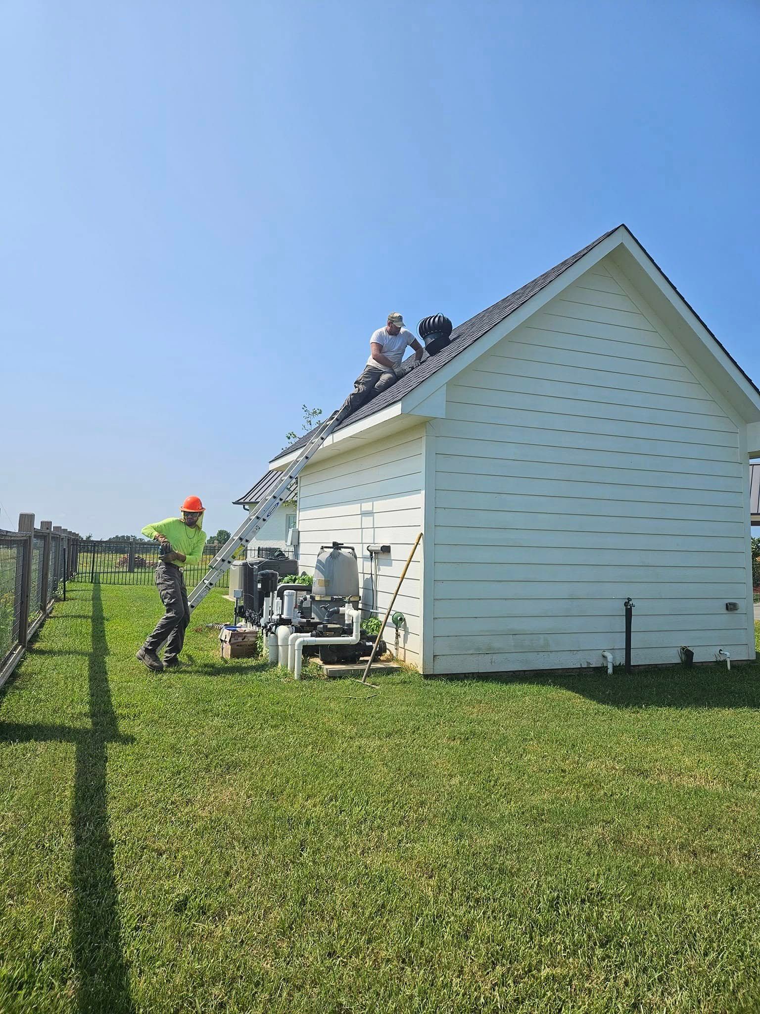 Two workers on a roof, one on a ladder. Green grass, white building, blue sky.
