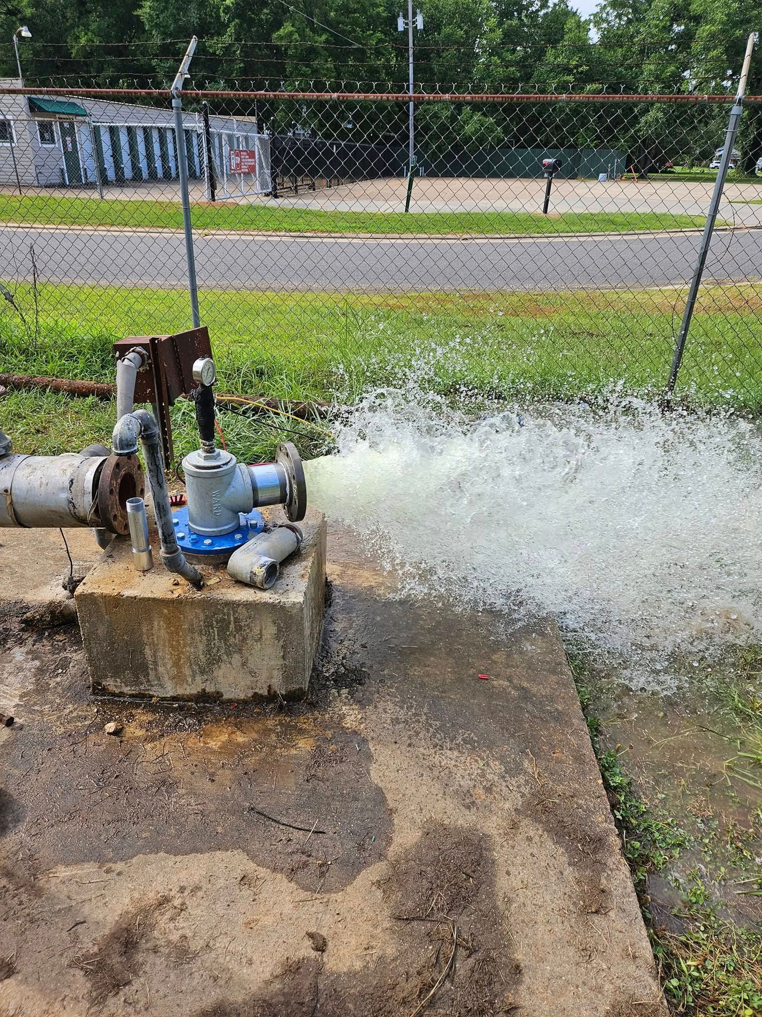 Water spraying from a blue valve attached to a concrete block next to a chain-link fence and grassy area.