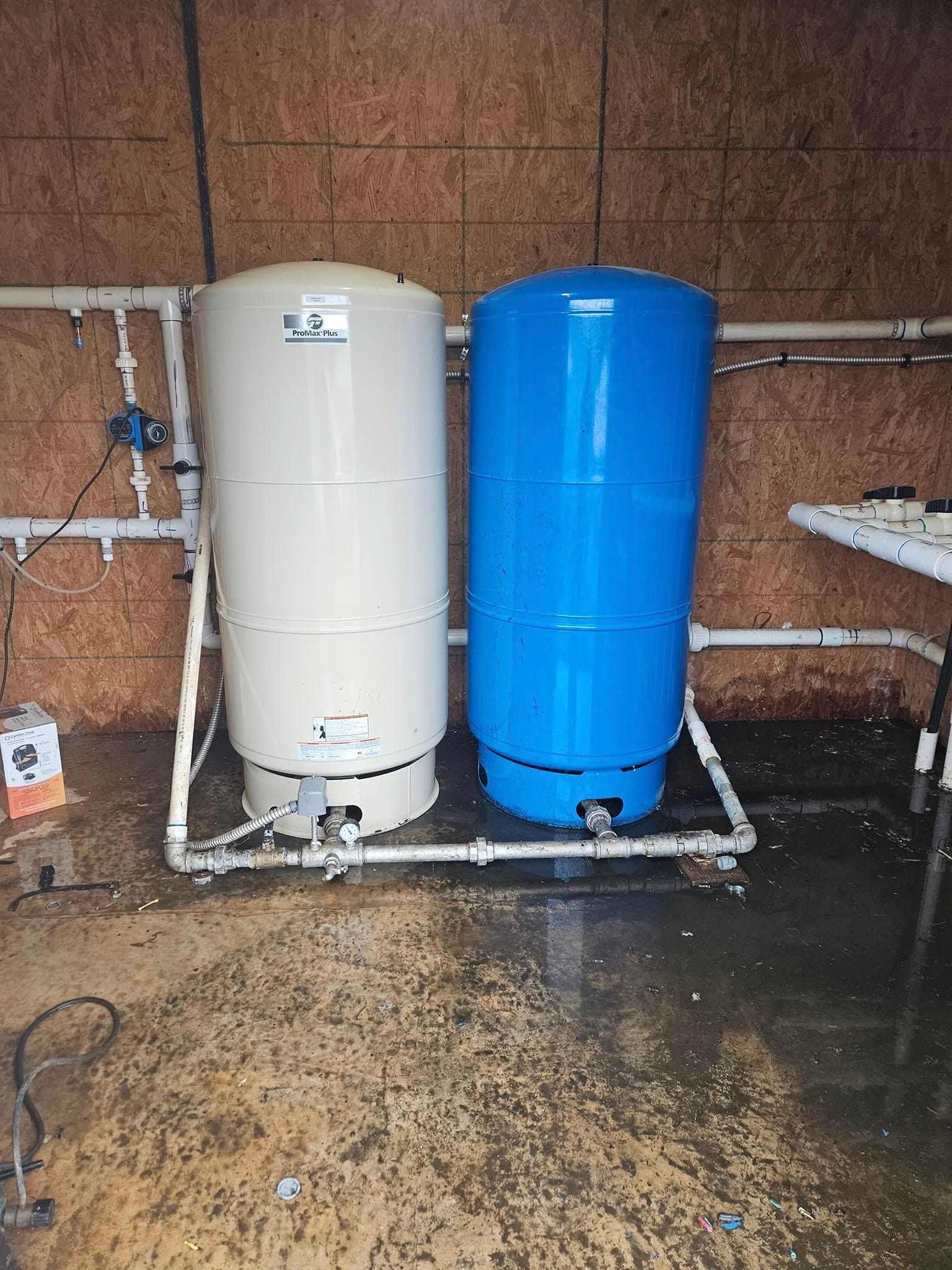 Two vertical water pressure tanks, tan and blue, on a PVC pipe frame, set in a room.