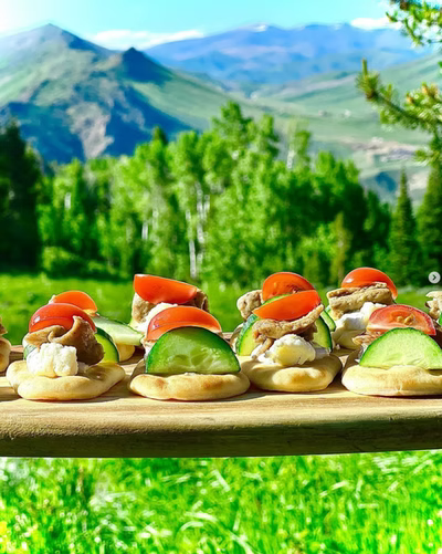 Small appetizers with cucumber, tomatoes, and toppings sit on a wooden board, with mountains and trees in the background.