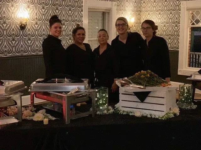 Five staff members in black attire stand behind a buffet table with food, decorations, and serving equipment. The setting is an event space.