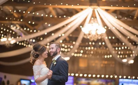 Bride and groom dancing at their wedding reception under string lights and draped fabric. They are gazing at each other in a ballroom.