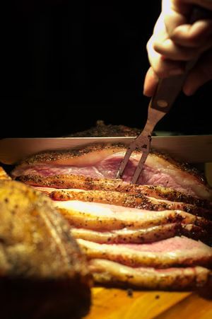 Close-up of someone carving a roast beef, showing pink slices and a fork. Dark background.