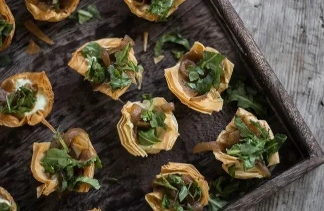Appetizer bites in golden, flaky pastry cups, filled with a mix of greens and what appear to be onions, arranged on a wooden tray.