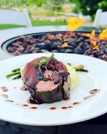 Plate of food with a dark, saucy meat roll, mashed potatoes, asparagus, and microgreens. In an outdoor setting, a fire pit is in the background.