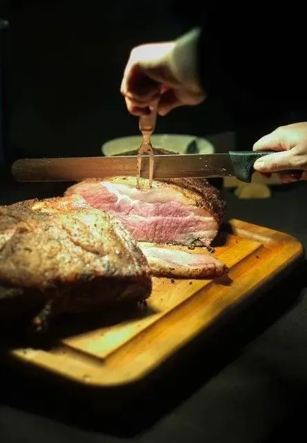 Person carving a cooked roast beef on a wooden cutting board with a carving fork and knife. The meat is pink inside.
