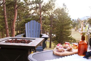 A cozy outdoor patio with a fire pit, blue Adirondack chair, and a wooden serving board with food, set against a forest.