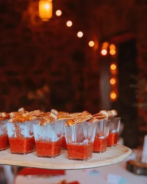 Tray of red soup shots topped with bread, served at an event with warm lighting and a brick wall background.