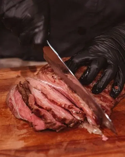 Person in black gloves slicing a cooked steak on a wooden cutting board, revealing pink interior and browned exterior.