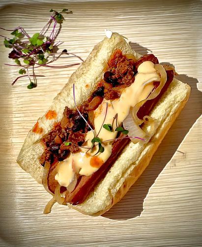 Hot dog in a bun with chili, onions, and sauce, garnished with microgreens, on a wooden tray.